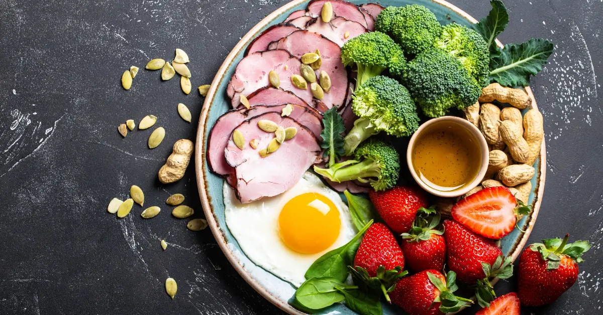 Prato saudável com ovos fritos na manteiga, fatias de carne, brocolis e folhas verdes temperadas com azeite extra virgem e sementes, acompanhado de morangos frescos.